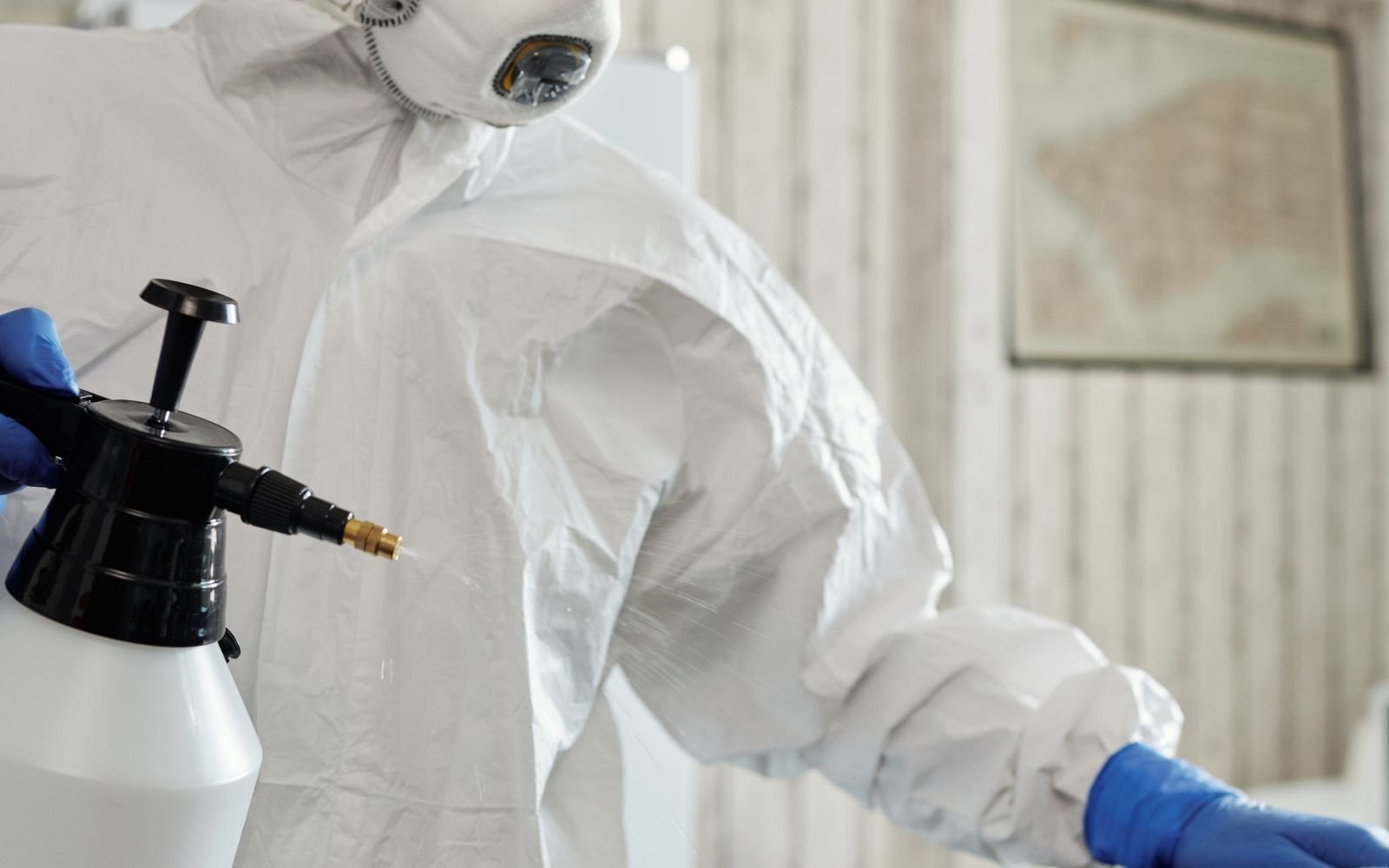 Intervenant en combinaison intégrale à usage unique, masque filtrant et gants nitrile, appliquant un produit au pulvérisateur dans une pièce.